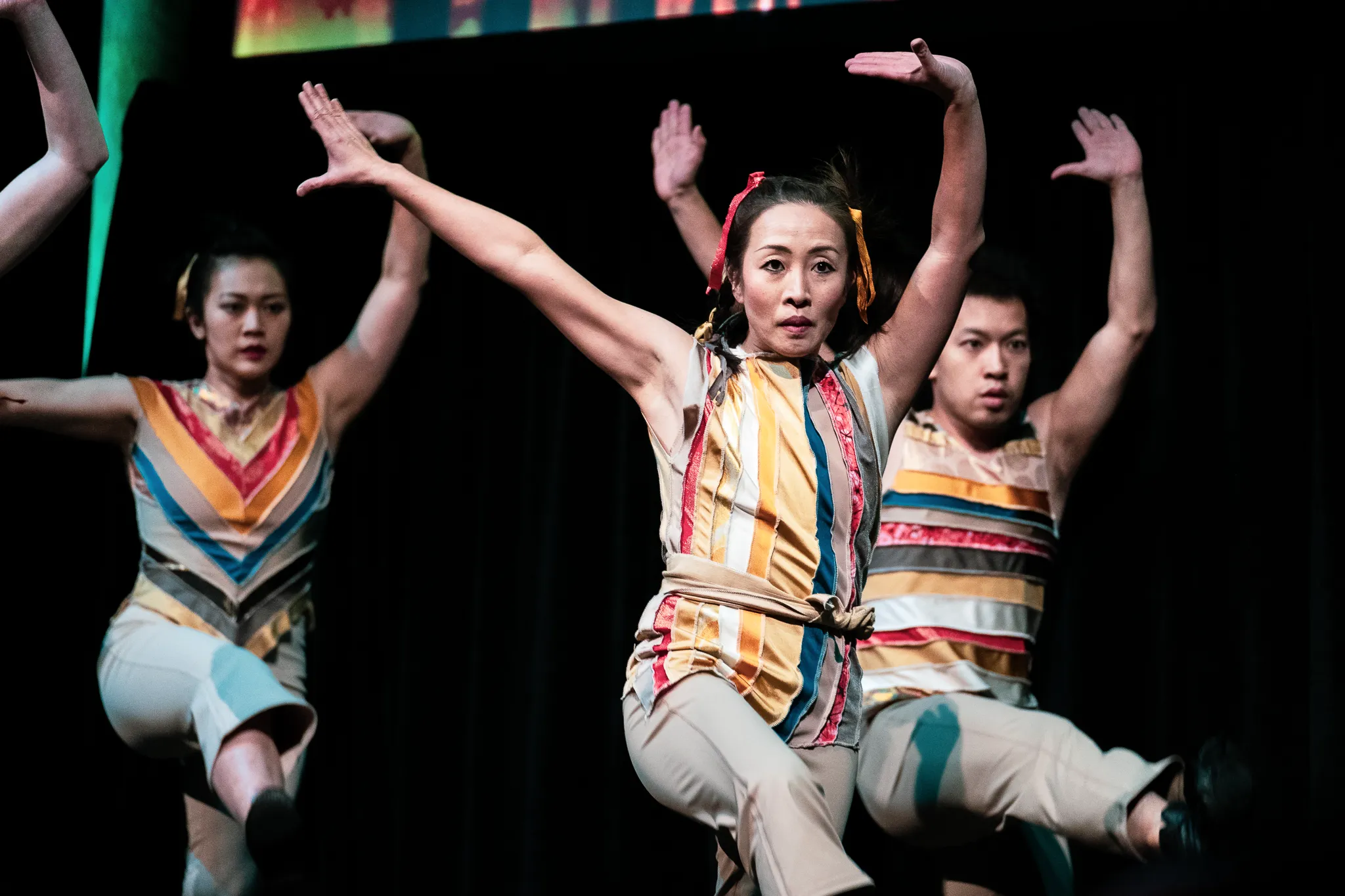 Dancers in striped costumes with raised arms and bent knees, led by a dancer looking toward the audience