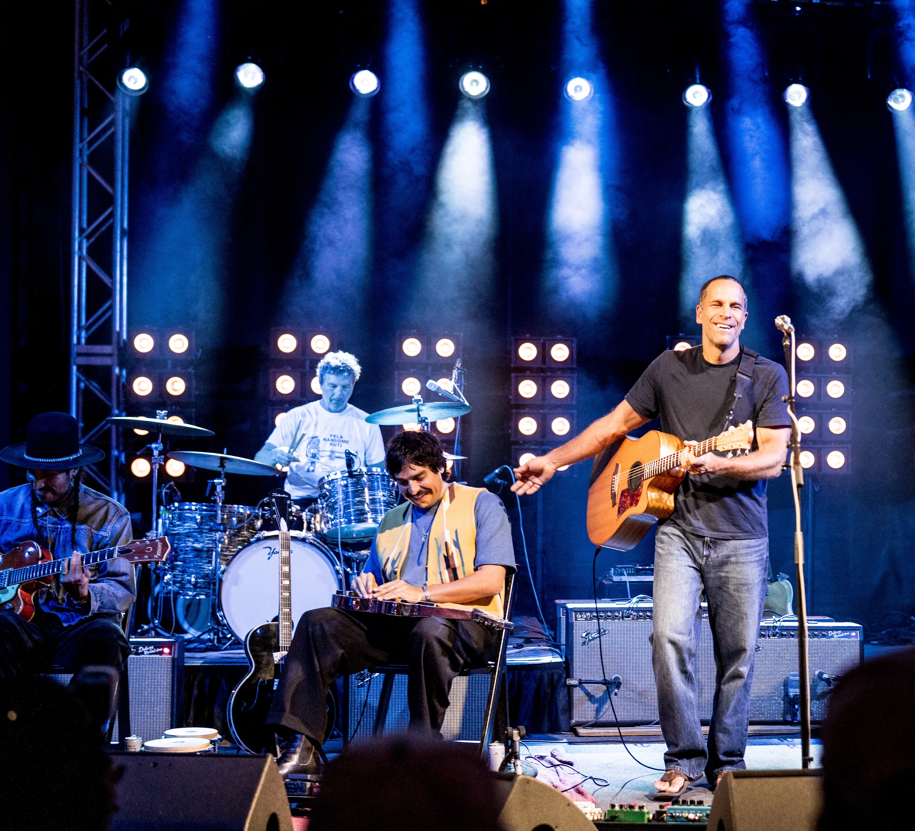 Jack Johnson smiling and gesturing toward a seated musician while holding his guitar