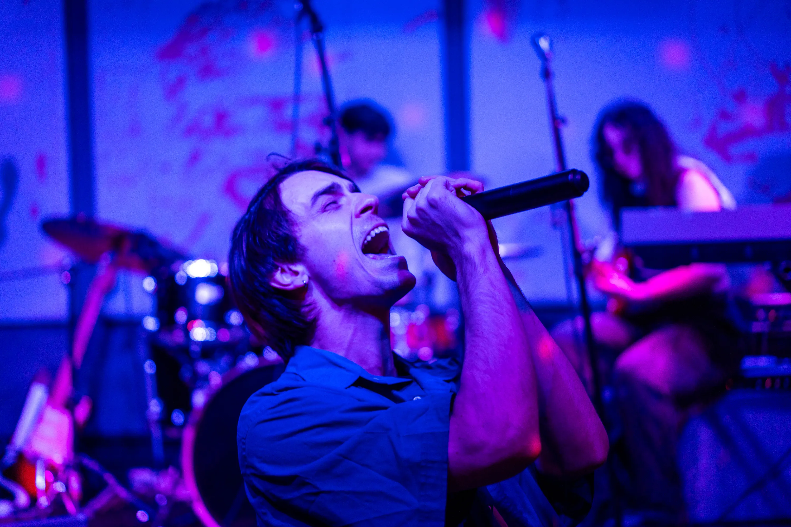 A vocalist leaning back into a handheld microphone, with the band behind him under blue and magenta lights