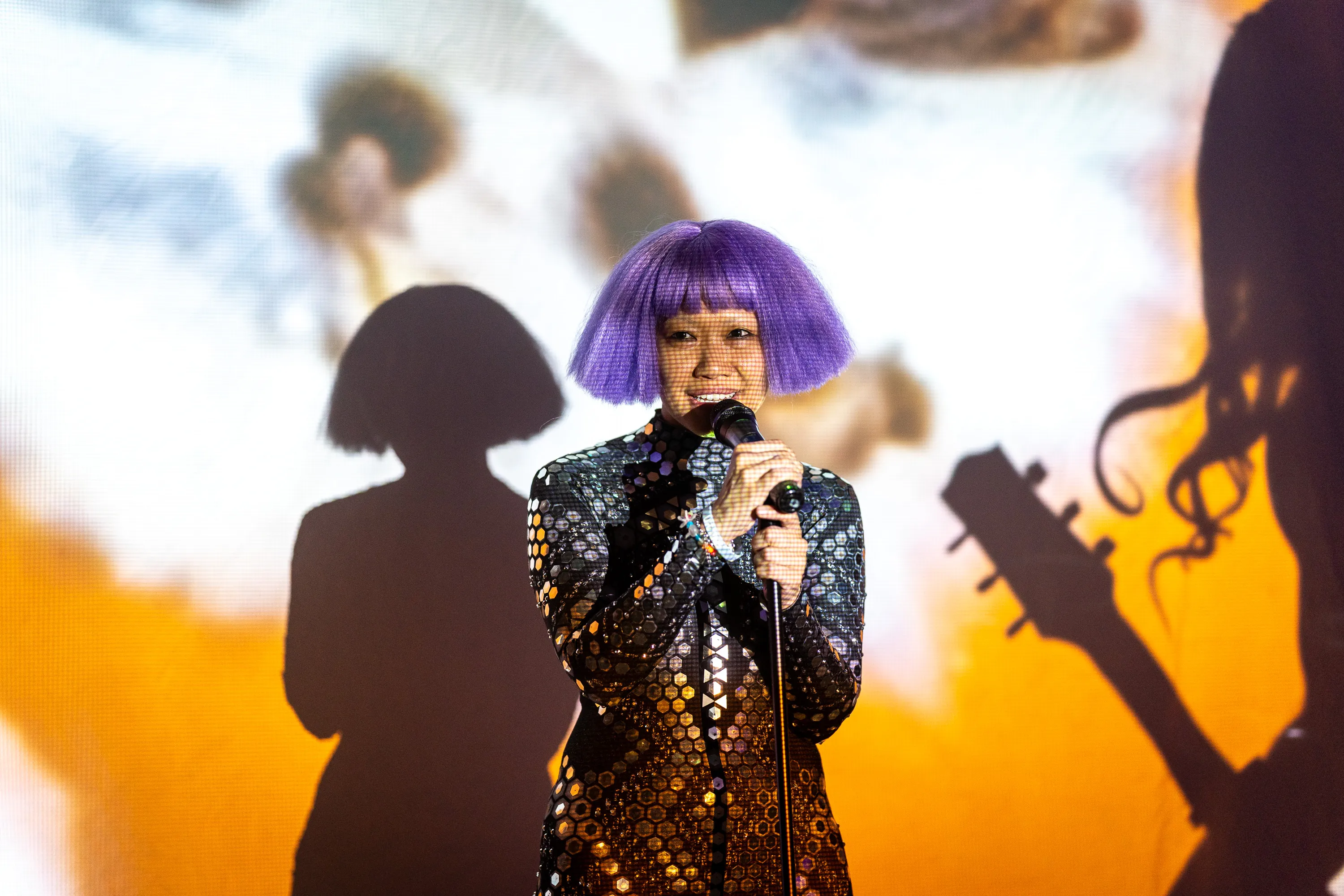 Olive singing in a mirrored outfit and purple bob, framed by projected shadows
